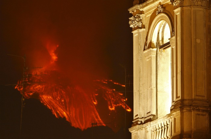 Imagem mostra a Catedral de Zafferana Etnea, com o vulcão Etna ao fundo, entrando em erupção